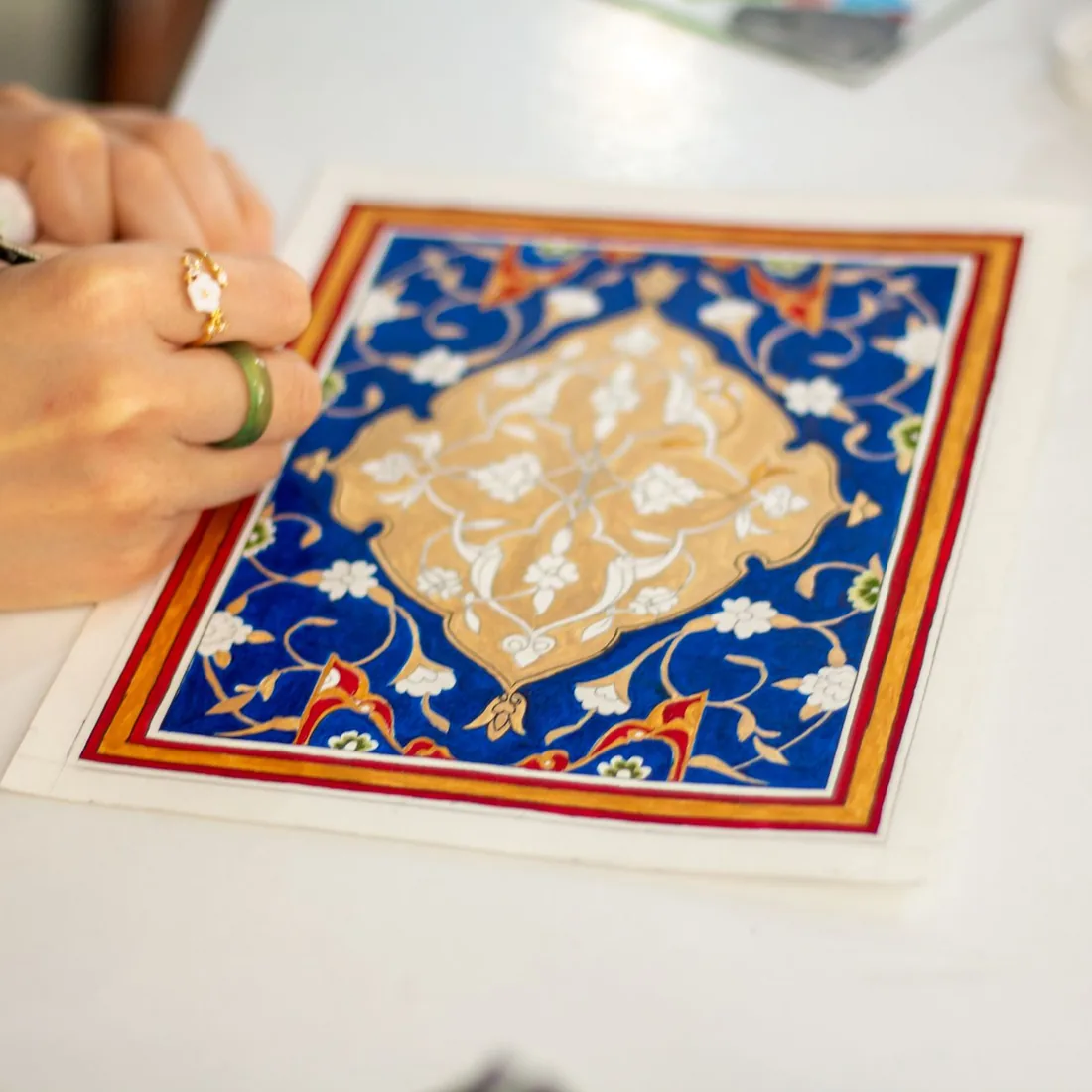 Mano sujetando un pincel fino sobre una miniatura ornamental pintada en azul, rojo y dorado